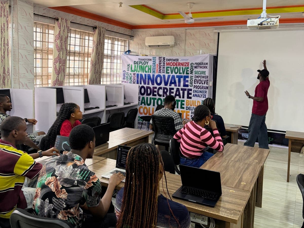 A trainer points at a projector screen while a group of learners with laptops watch from wooden tables in a training room.