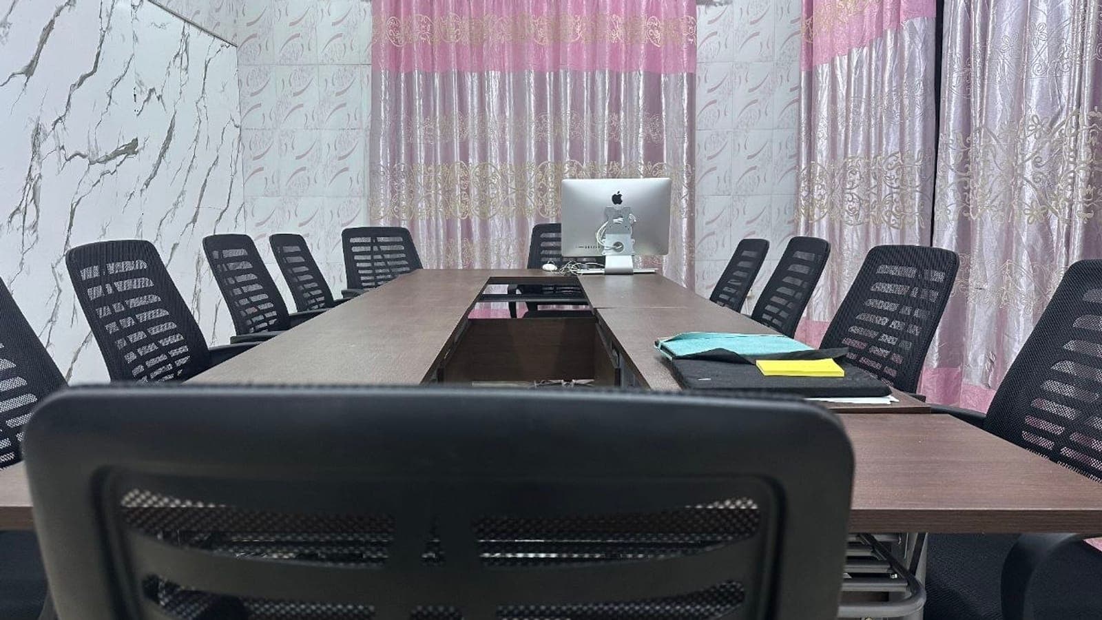 A meeting room with a long wooden table, black mesh office chairs and a desktop computer at the far end, in front of pink curtains and a marble-effect wall.